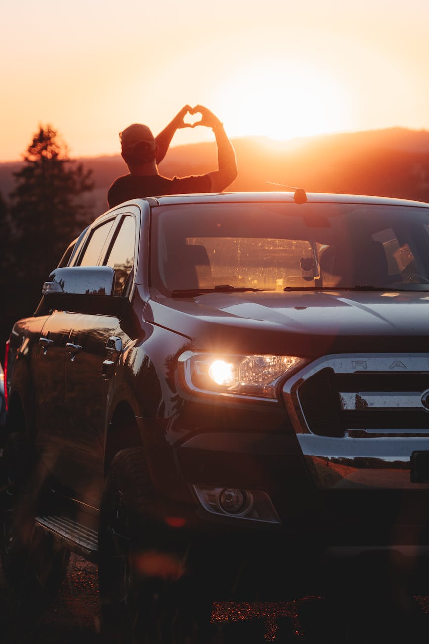 person standing in the back of a pick up truck making a heart gesture towards the setting sun